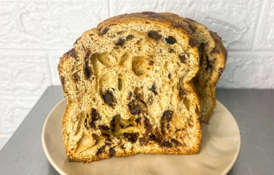 Fotografia em tons claros com um panettone de chocolate artesanal ao centro. O panettone possui massa aerada várias gotas de chocolate