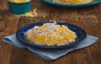 Imagem aproximada da receita de Cuscuz em tom amarelo, em um prato azul escuro, sobre um tecido em tom de azul claro. O cuscuz está decorado com coco ralado e, ao fundo, estão um pote com fubá amarelo e um outro prato com outro cuscuz