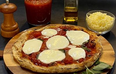 Foto da receita de pizza napolitana servida sobre uma tábua de madeira redonda com folhas manjericão ao lado. Ao fundo, há um bowl de vidro com muçarela, um vidro de azeite, um moedor de sal e um vidro de molho pomodoro Foto da receita de pizza napolitana servida sobre uma tábua de madeira redonda com folhas manjericão ao lado. Ao fundo, há um bowl de vidro com muçarela, um vidro de azeite, um moedor de sal e um vidro de molho pomodoro