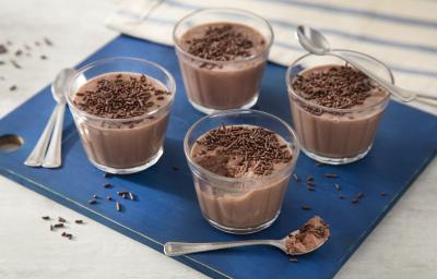 Fotografia em tons de azul em uma bancada de madeira clara, uma tábua azul quadrada com quatro copinhos de vidro com a mousse de chocolate dentro e decoradas com os chocolates granulados. Fotografia em tons de azul em uma bancada de madeira clara, uma tábua azul quadrada com quatro copinhos de vidro com a mousse de chocolate dentro e decoradas com os chocolates granulados.