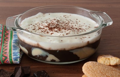 Em cima de uma mesa, recipiente redondo de vidro com creme de chocolate, decorado com chantilly e raspas de chocolate.