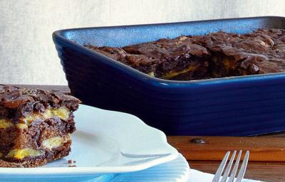 Fotografia de um pedaço da torta de banana em camadas em cima de um prato redondo e azul com um garfo ao lado, ao lado refratário azul com o restante da torta, tudo sobre bancada de madeira e fundo cinza. Fotografia de um pedaço da torta de banana em camadas em cima de um prato redondo e azul com um garfo ao lado, ao lado refratário azul com o restante da torta, tudo sobre bancada de madeira e fundo cinza.