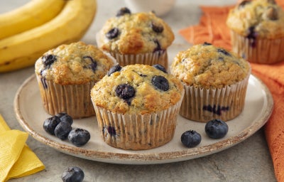 Imagem mostra quatro muffins dourados com pedaços de mirtilos azuis. Estão em forminhas brancas sobre um prato branco.