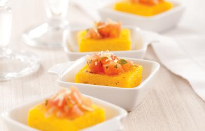 Fotografia em tons de laranja em uma bancada de madeira clara, um pano bege, quatro potinhos brancos pequenos com a polenta com tomate dentro de cada um. Fotografia em tons de laranja em uma bancada de madeira clara, um pano bege, quatro potinhos brancos pequenos com a polenta com tomate dentro de cada um.