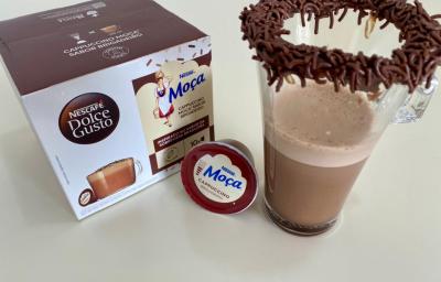 Fotografia de uma xícara de vidro com uma bebida de dolce gusto brigadeiro, com brigadeiro e granulado na borda. Ao lado tem uma cápsula de dolce gusto brigadeiro e a caixa com as cápsulas. Fotografia de uma xícara de vidro com uma bebida de dolce gusto brigadeiro, com brigadeiro e granulado na borda. Ao lado tem uma cápsula de dolce gusto brigadeiro e a caixa com as cápsulas.