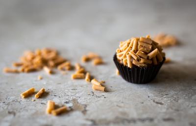 fotografia em tons de cinza, marrom e preto de uma bancada cinza vista de frente. Contém uma forminha preta com um brigadeiro dentro, por cima raspas de caramelo e na bancada contém as raspas espalhadas.
