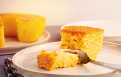Fotografia de um prato grande de vidro com uma fatia grande de bolo de milho e um garfo cortando um pedaço. No fundo tem o bolo inteiro, sem essa fatia, sobre um apoio raso de cor clara. Fotografia de um prato grande de vidro com uma fatia grande de bolo de milho e um garfo cortando um pedaço. No fundo tem o bolo inteiro, sem essa fatia, sobre um apoio raso de cor clara.