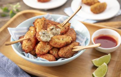 Fotografia em tons de bege em uma bancada de madeira clara, uma tábua de madeira, um pano azul, um prato redondo fundo com vários bolinhos de peixe, um potinho branco pequeno com molho de pimenta e pedaços de limão. Ao fundo, pratinhos brancos com bolinho Fotografia em tons de bege em uma bancada de madeira clara, uma tábua de madeira, um pano azul, um prato redondo fundo com vários bolinhos de peixe, um potinho branco pequeno com molho de pimenta e pedaços de limão. Ao fundo, pratinhos brancos com bolinho