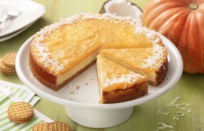 Fotografia em tons de verde e laranja de uma bancada de madeira verde com um paninho listrado verde e branco com biscoitos  e uma bailarina branca com o cheesecake de abóbora. Ao fundo uma abóbora, um coco e um prato quadrado branco.