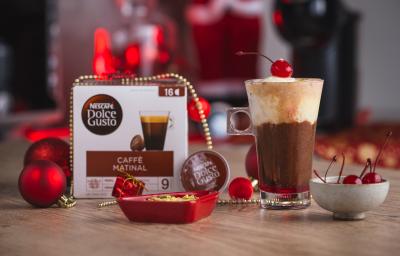 Fotografia em tons de vermelho em uma bancada de madeira, uma xícara alta com o affogato natalino decorado com uma cereja. Ao fundo, enfeites e decoração de natal.