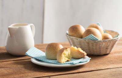 Fotografia em tons de azul em uma bancada de madeira com um prato branco raso e um guardanapo de papel azul com bolinhas brancas e o pãozinho de ninho em cima e cortado ao meio. Ao lado, uma cesta de vime com mais pãezinhos de ninho.