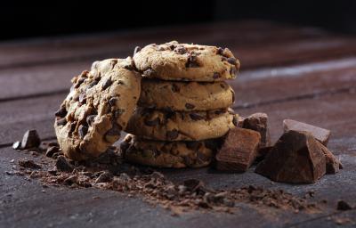 Fotografia de quatro cookies com gotas de chocolate, um sobre o outro. Encostado neles tem mais um cookie, ao lado de pedaços de chocolate, sobre uma mesa de madeira escura. Fotografia de quatro cookies com gotas de chocolate, um sobre o outro. Encostado neles tem mais um cookie, ao lado de pedaços de chocolate, sobre uma mesa de madeira escura.