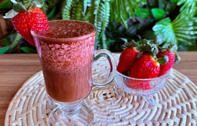 Fotografia em tons de rosa e marrom com uma caneca grande ao centro. Dentro da caneca existe uma bebida gelada a base de cápsulas de Dolce Gusto, e morangos congelados. Ao lado, existe um potinho com vários morangos. Fotografia em tons de rosa e marrom com uma caneca grande ao centro. Dentro da caneca existe uma bebida gelada a base de cápsulas de Dolce Gusto, e morangos congelados. Ao lado, existe um potinho com vários morangos.