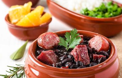 Fotografia de um recipiente médio marrom clássico com uma feijoada com caldo de bacon, linguiça e bacon com uma folha de salsinha. Ao fundo tem outros recipientes iguais, porém menores, com laranja, arroz, couve e farofa. Fotografia de um recipiente médio marrom clássico com uma feijoada com caldo de bacon, linguiça e bacon com uma folha de salsinha. Ao fundo tem outros recipientes iguais, porém menores, com laranja, arroz, couve e farofa.