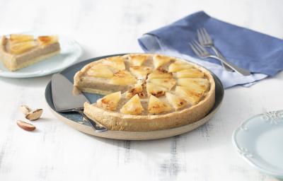Fotografia em tons de azul em uma bancada de madeira branca, um pano azul jeans, um prato redondo raso grande com a torta de castanha-do-pará em cima dele. Ao lado, um prato pequeno com uma fatia da torta.