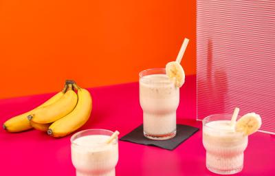 Fotografia de três copos de vidro com vitamina de banana e goiaba, e em cada copo, tem um canudo e uma rodela de banana decorando-o. Ao lado esquerdo, um cacho com quatro bananas, sobre uma mesa rosa com fundo laranja.