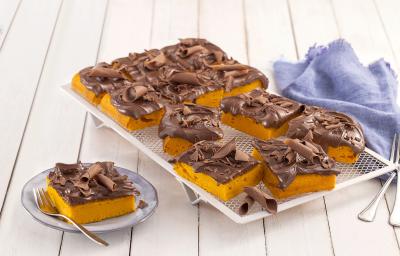 Fotografia em tons de azul em uma bancada de madeira branca, um pano azul, um apoiador de grelha branco com vários pedaços do bolo de cenoura com cobertura de chocolate. Ao lado, um prato pequeno com uma fatia do bolo.