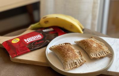 Fotografia em tons de marrom em uma bancada de madeira com uma tábua de madeira e ao centro, um prato redondo com dois pasteis. Ao lado, um tablete de chocolate e um cacho de bananas ao fundo. Fotografia em tons de marrom em uma bancada de madeira com uma tábua de madeira e ao centro, um prato redondo com dois pasteis. Ao lado, um tablete de chocolate e um cacho de bananas ao fundo.