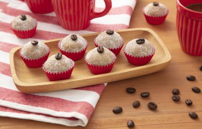 Fotografia em tons de vermelho de uma bancada de madeira, ao centro um pano listrado vermelho e branco e uma bandeja de madeira com seis brigadeiros em forminhas vermelhas, em cima de cada um deles um grão de café. Fotografia em tons de vermelho de uma bancada de madeira, ao centro um pano listrado vermelho e branco e uma bandeja de madeira com seis brigadeiros em forminhas vermelhas, em cima de cada um deles um grão de café.