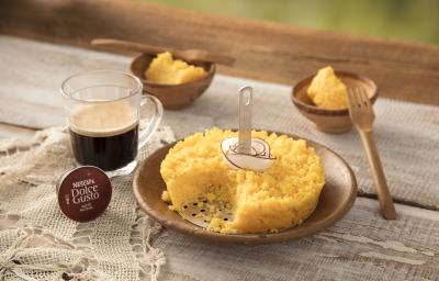 Fotografia em tons de amarelo, branco e marrom de uma bancada com um pano rendado, uma cápsula de Dolce Gusto e uma xícara transparente com café. Ao lado um prato pequeno redondo com o cuscuz e tiras de coco. Ao fundo, dois potinhos com cuscuz e 2 garfos.
