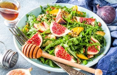 Em cima de uma mesa, prato com salada com mix de folhas, frutas em cubos e figo.