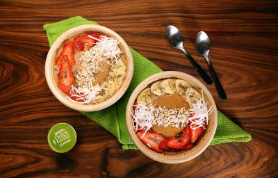 Fotografia em tons de verde em uma bancada de madeira escura, um pano verde, dois bowls bege com morango, banana e lascas de coco e duas colheres ao lado. Fotografia em tons de verde em uma bancada de madeira escura, um pano verde, dois bowls bege com morango, banana e lascas de coco e duas colheres ao lado.