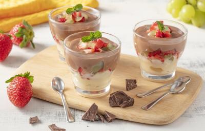 Fotografia em tons de amarelo em uma bancada cinza com uma tábua de madeira no centro e três potinhos de vidro com o bombom de chocolate, creme e frutas. Ao lado, pedaços de chocolate, morangos e um cacho de uvas espalhados.