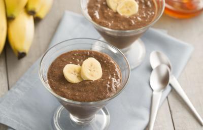 Fotografia em tons de cinza, marrom, amarelo e azul de uma bancada cinza vista de cima, ao centro um pano azul com duas taças transparente ambas contém mousse de chocolate e duas fatias de banana. Ao fundo um cacho de banana, mel e ao lado duas colheres