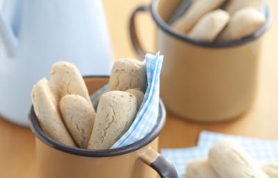Fotografia em tons de azul em uma mesa de madeira com um paninho azul claro com bolinhas brancas, ao centro uma xícara bege com os biscoitinhos nutritivos. Ao fundo, uma jarra azul claro. Fotografia em tons de azul em uma mesa de madeira com um paninho azul claro com bolinhas brancas, ao centro uma xícara bege com os biscoitinhos nutritivos. Ao fundo, uma jarra azul claro.