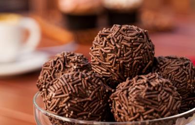 Fotografia de vários brigadeiros enrolados no granulado e recheados com panettone. Os brigadeiros estão em um recipiente fundo de vidro, mas que só aparece na borda, e está sobre uma mesa de madeira.