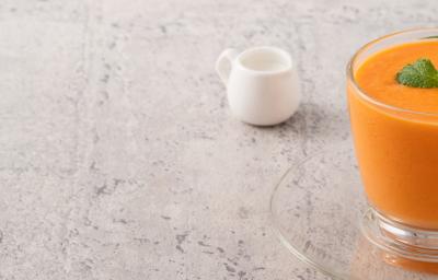 Fotografia em tons claros de uma vitamina laranja com folhas de hortelã por cima em um copo de vidro, apoiado sobre um prato pequeno de vidro. Ao fundo, um pote pequeno branco de vidro, sobre uma mesa em tons de cinza. Fotografia em tons claros de uma vitamina laranja com folhas de hortelã por cima em um copo de vidro, apoiado sobre um prato pequeno de vidro. Ao fundo, um pote pequeno branco de vidro, sobre uma mesa em tons de cinza.