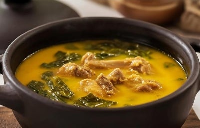 Receita de frango servido no caldo de tucupi com folhas de jambu. Uma foto bem aproximada que mostra uma panela de barro preta com tudo dentro.