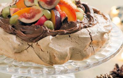 fotografia tirada de frente, contém um suporte para servir sobremesas transparente por cima um merengue coberto de chocolate com frutas para decorar.