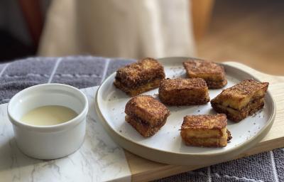Foto aproximada de um prato com pedaços de Pão com Canela French Toast, ao lado de um potinho pequeno com um pouco de Leite Moça, tudo sobre uma toalha cinza quadriculada