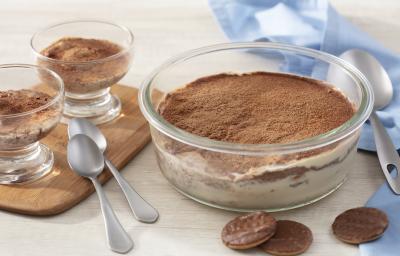 Fotografia em tons de azul em uma bancada de madeira clara, um paninho azul, uma tábua de madeira, um recipiente de vidro redondo com o tiramisú feito com calipso. Ao lado, em cima da tábua, duas taças com o tiramisú entro. Fotografia em tons de azul em uma bancada de madeira clara, um paninho azul, uma tábua de madeira, um recipiente de vidro redondo com o tiramisú feito com calipso. Ao lado, em cima da tábua, duas taças com o tiramisú entro.