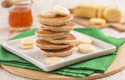 Fotografia em tons de verde em uma bancada de madeira clara, uma tábua de madeira redonda com um pano verde em cima, um prato quadrado branco e seis panquequinhas empilhadas com rodelas de banana em torno delas. Ao fundo, um potinho de mel e uma banana. Fotografia em tons de verde em uma bancada de madeira clara, uma tábua de madeira redonda com um pano verde em cima, um prato quadrado branco e seis panquequinhas empilhadas com rodelas de banana em torno delas. Ao fundo, um potinho de mel e uma banana.