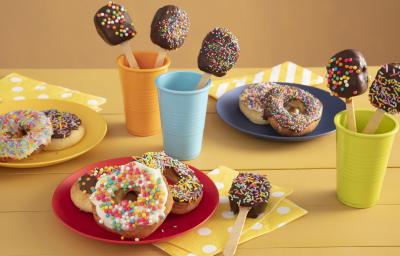 Fotografia em tons de amarelo em uma bancada de madeira amarela com três copos coloridos, azul, laranja e verde com pirulitos de chocolate dentro dele. Ao centro, um prato vermelho com três donuts cobertos com granulados coloridos. Fotografia em tons de amarelo em uma bancada de madeira amarela com três copos coloridos, azul, laranja e verde com pirulitos de chocolate dentro dele. Ao centro, um prato vermelho com três donuts cobertos com granulados coloridos.