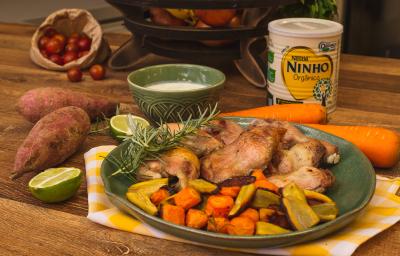 Fotografia em tons de marrom e verde de uma bancada de madeira, paninho xadrez amarelo e branco e um prato verde com pedaços de frango, cenoura e batata-doce. Ao fundo lata de NINHO e potinho com molho. Fotografia em tons de marrom e verde de uma bancada de madeira, paninho xadrez amarelo e branco e um prato verde com pedaços de frango, cenoura e batata-doce. Ao fundo lata de NINHO e potinho com molho.
