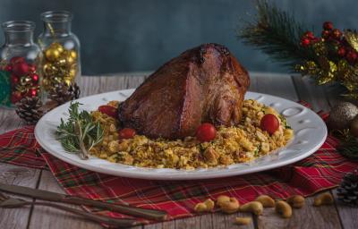 Foto da receita de pernil com farofa crocante, servido em um prato branco redondo, sobre uma mesa de madeira toda decorada com itens natalinos. Há algumas castanhas de caju espalhadas na mesa e um conjunto de talheres à frente do prato Foto da receita de pernil com farofa crocante, servido em um prato branco redondo, sobre uma mesa de madeira toda decorada com itens natalinos. Há algumas castanhas de caju espalhadas na mesa e um conjunto de talheres à frente do prato