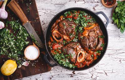 Fotografia vista de cima uma panela de ferro com ossobucos com molho e salsinha por cima. Ao lado, uma tábua de madeira em tom escuro, e em cima, uma faca, salsinha picada, duas cabeças de alho, sal grosso e um limão siciliano sem a casca. Fotografia vista de cima uma panela de ferro com ossobucos com molho e salsinha por cima. Ao lado, uma tábua de madeira em tom escuro, e em cima, uma faca, salsinha picada, duas cabeças de alho, sal grosso e um limão siciliano sem a casca.