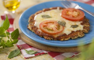 fotografia em tons de azul, vermelho e marrom de uma bancada marrom vista de cima, contém uma tábua redonda, um prato redondo azul com uma mini pizza coberta com queijo, duas fatias de tomate e manjericão e ao lado um garfo para servir.