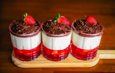 Fotografia em tons de marrom com três potes de vidro ao centro. Dentro de cada pote existe uma sobremesa em camadas feita com gelatina de morango, creme branco e coberta com ganache de chocolate. Fotografia em tons de marrom com três potes de vidro ao centro. Dentro de cada pote existe uma sobremesa em camadas feita com gelatina de morango, creme branco e coberta com ganache de chocolate.