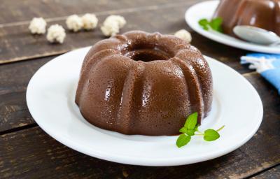 Fotografia de um pudim pequeno de chocolate meio amargo e folhas de hortelã que estão em cima de um prato raso de vidro na cor branco. Ao fundo, o mesmo pudim sobre um prato, um pano azul e algumas pipocas sobre uma mesa de madeira de tom escuro. Fotografia de um pudim pequeno de chocolate meio amargo e folhas de hortelã que estão em cima de um prato raso de vidro na cor branco. Ao fundo, o mesmo pudim sobre um prato, um pano azul e algumas pipocas sobre uma mesa de madeira de tom escuro.