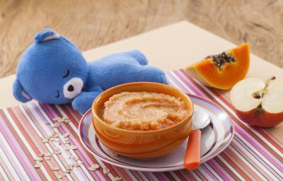 Fotografia em tons de laranja e azul de uma bancada de madeira com um paninho listrado colorido, um potinho laranja com o purêzinho de mamão, atrás um ursinho de pelúcia azul. Ao fundo, meio pedaço de maçã e meio pedaço de mamão.