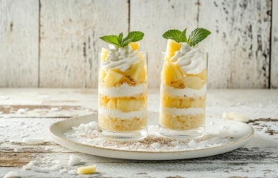 Fotografia em tons claros de dois copos longos com a sobremesa em camadas com pedaços de abacaxi, decorados com chantilly e hortelã.