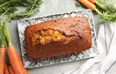 Bolo de cenoura retangular, dourado e rústico, com superfície rachada e pedaços de cenoura visíveis. Está sobre prato preto e branco, cercado por cenouras frescas e ao lado de um pano branco com listras cinza. Bolo de cenoura retangular, dourado e rústico, com superfície rachada e pedaços de cenoura visíveis. Está sobre prato preto e branco, cercado por cenouras frescas e ao lado de um pano branco com listras cinza.