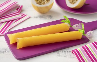 Fotografia em tons de branco e roxo de uma bancada de madeira branca e uma tábua roxa, sobre ela dois geladinhos de maracujá. Ao lado paninhos listrados em tons de roxo e branco, ao fundo um prato redondo roxo com um maracujá cortado ao meio.