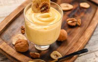 Fotografia de uma flan de nozes e leite MOÇA com uma rodela de banana e uma noz por cima dentro de um recipiente de vidro decorativo. O creme está sobre uma tábua de madeira com várias nozes espalhadas por cima, juntamente com uma colher de sobremesa. Fotografia de uma flan de nozes e leite MOÇA com uma rodela de banana e uma noz por cima dentro de um recipiente de vidro decorativo. O creme está sobre uma tábua de madeira com várias nozes espalhadas por cima, juntamente com uma colher de sobremesa.