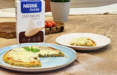 Fotografia em tons de marrom com um prato azul ao centro. Em cima do prato existe uma pizza com massa de Aveia coberta com queijo muçarela e orégano. Ao fundo existe uma embalagem de Farelo de Aveia NESTLÉ.