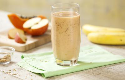 Fotografia em tons de rosa, verde e branco, com copo ao centro com vitamina sobre guardanapo verde, no entorno duas bananas, maçã cortada em pedaços e um pedaço de mamão sobre peça de madeira, potinho com linhaça, tudo sobre bancada em tons de branco. Fotografia em tons de rosa, verde e branco, com copo ao centro com vitamina sobre guardanapo verde, no entorno duas bananas, maçã cortada em pedaços e um pedaço de mamão sobre peça de madeira, potinho com linhaça, tudo sobre bancada em tons de branco.